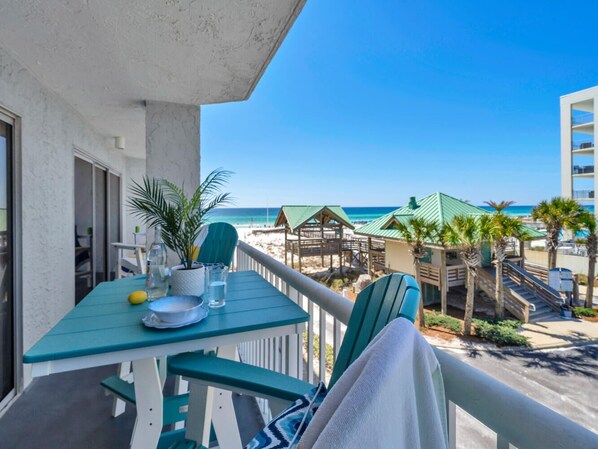 Un balcón con una mesa dispuesta para dos, con vista a una playa con palmeras y una estructura de madera.