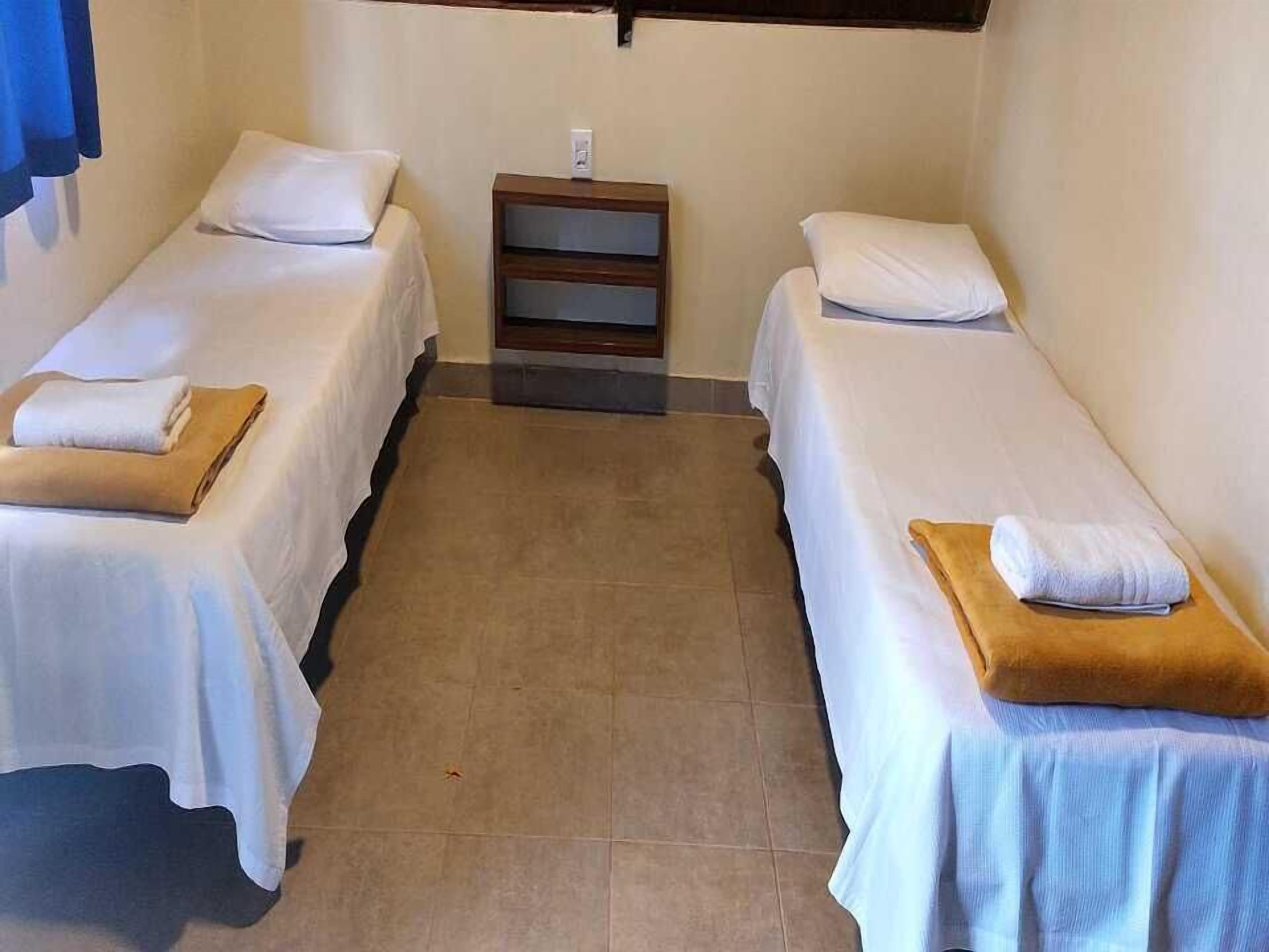 Ein Hotelzimmer mit zwei Einzelbetten, jeweils mit Kissen und einem gefalteten Handtuch. Zwischen den Betten befindet sich ein Nachttisch aus Holz mit einem kleinen Gegenstand darauf.