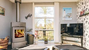 A living room with a wood-burning stove, a television, and a large window with a view of trees and buildings. - 4 Person Holiday Home in Vordingborg (Vordingborg)
