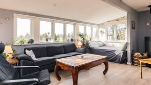 A spacious living room with a dark grey sectional sofa, a wooden coffee table, and a black armchair. Large windows provide ample natural light and views of the outdoors. - 4 Person Holiday Home in Vordingborg (Vordingborg)