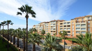 Een meerlaags hotel met een rij palmbomen ervoor, een voetpad en een helderblauwe lucht.