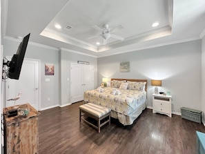 A bedroom with a bed, bedside tables, a bench, a ceiling fan, and a TV mounted on the wall.