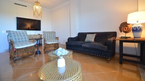 A living room with a TV, two patterned armchairs, a dark sofa, a glass coffee table with a vase, and a lamp on a side table. - San Bruno 73 - Two Bedroom Apartment, Sleeps 5 (Punta del moral)