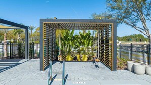 Un espacio de estar al aire libre cubierto, con estructura metálica y listones de madera, rodeado de plantas en macetas y un camino pavimentado.