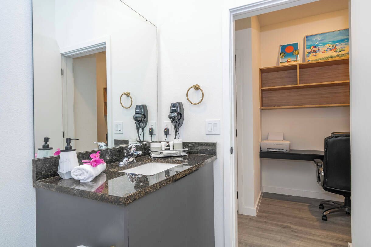 Une salle de bain moderne avec un comptoir en granit, un grand miroir et une serviette ornée de fleurs roses. On y trouve deux sèche-cheveux fixés au mur, une étagère avec une imprimante et un bureau avec une chaise.