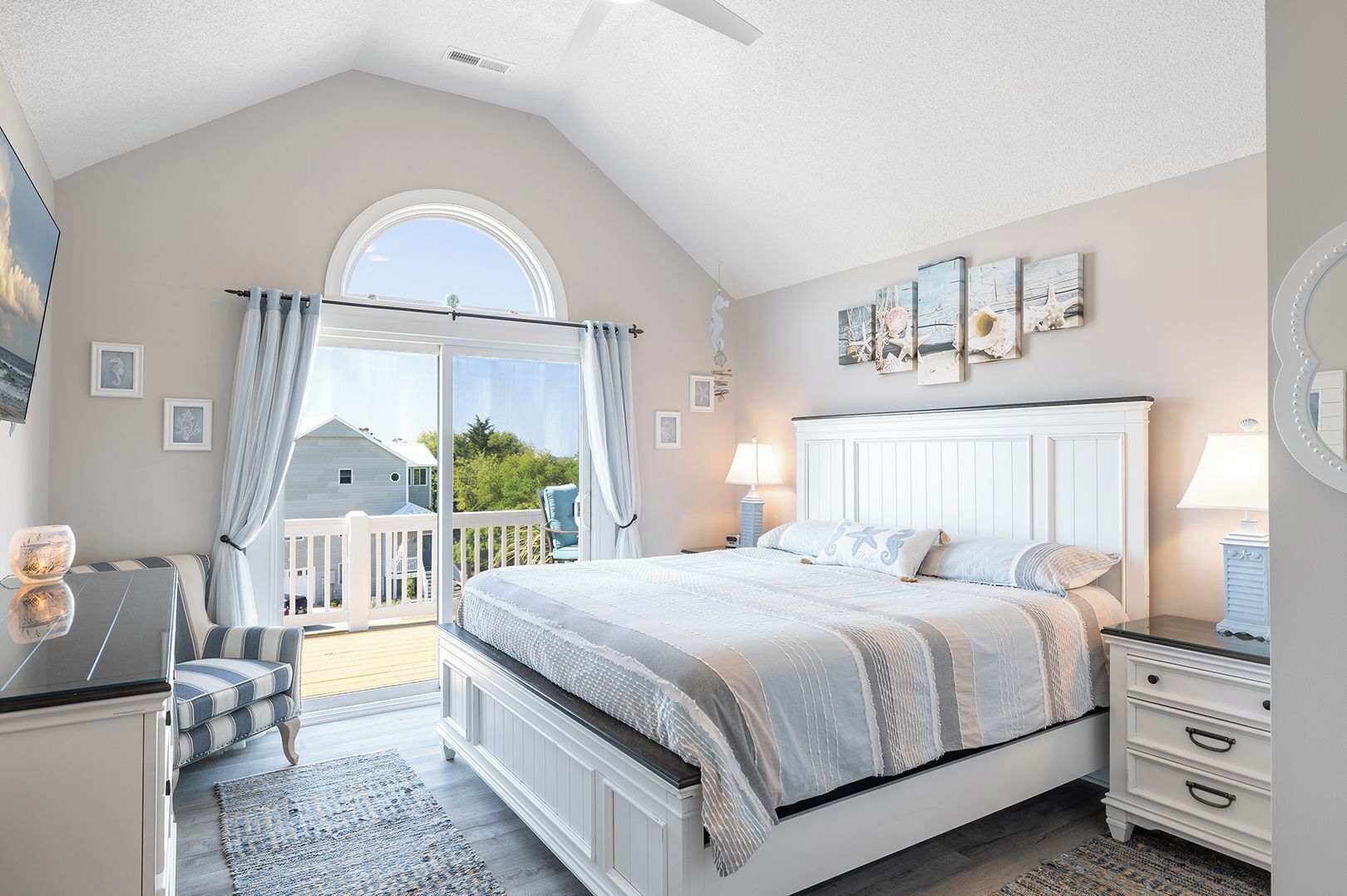Ein Schlafzimmer mit einem großen Bett, einem Nachttisch, einer Kommode, einem Sessel und einem Balkon mit Blick auf ein Haus und Bäume.