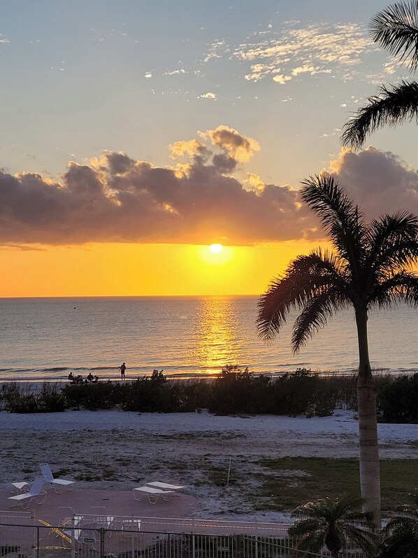 A sunset over the ocean with palm trees and beach chairs visible. - Seaside Serenity at EBT 105C (Fort Myers Beach)