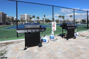 Two barbecue grills on a patio with a tennis court in the background. - Seaside Serenity at EBT 105C (Fort Myers Beach)