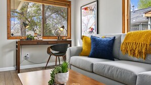 A living room with a desk, a large window, a grey sofa, and a yellow throw blanket. - Old Town Basecamp | 6 Blocks to Old Town Square (Fort Collins)