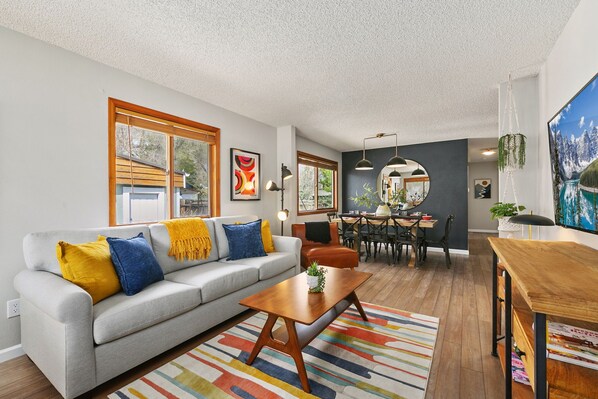 A modern living room with a gray sofa, wooden coffee table, and colorful rug. There is a dining area with a table and chairs, and a large painting on the wall.