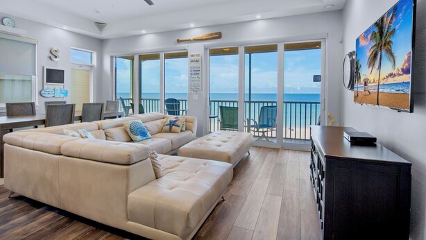 A spacious living room with a large beige sectional sofa, a ceiling fan, and a view of the ocean through large sliding glass doors. - Beachfront Stay! BBQ, Lanai and Cozy Beds! (Bradenton Beach)