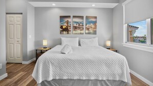 A bedroom with a large bed, two bedside tables with lamps, a cityscape artwork on the wall, and a window with a view of buildings. - Beachfront Stay! BBQ, Lanai and Cozy Beds! (Bradenton Beach)