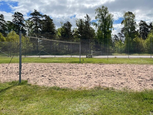 Una cancha de voleibol de arena, con una red, rodeada por una cerca de malla metálica y árboles.