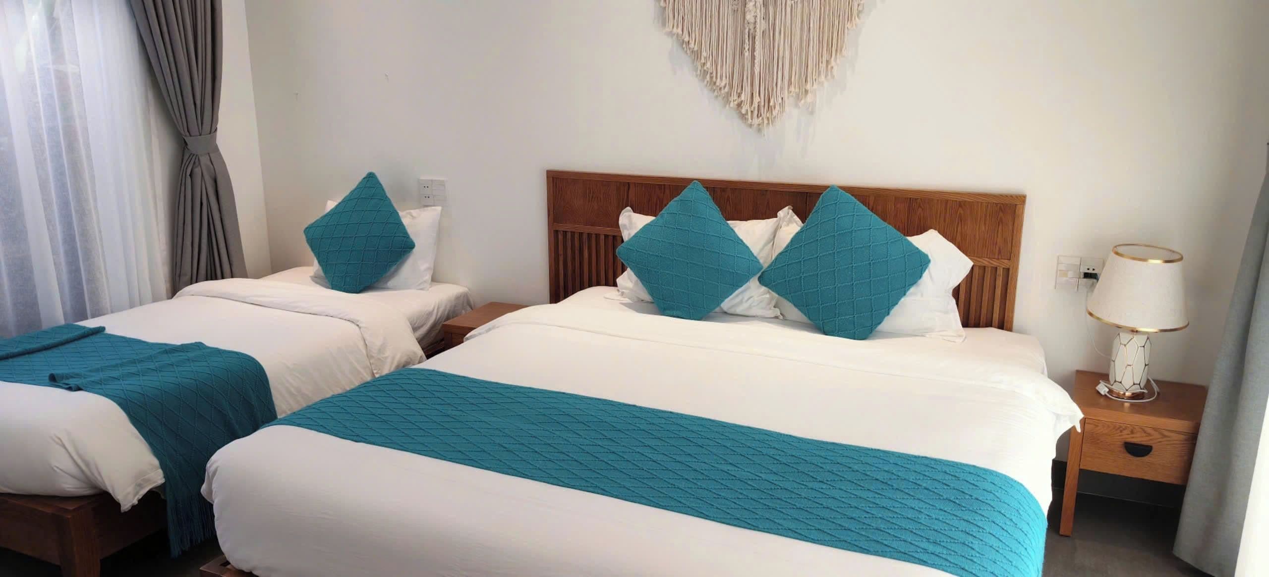 A hotel room with two beds, each with a white bedspread and teal pillows. There is a wooden headboard, a wall-mounted macrame piece, and a bedside table with a lamp.