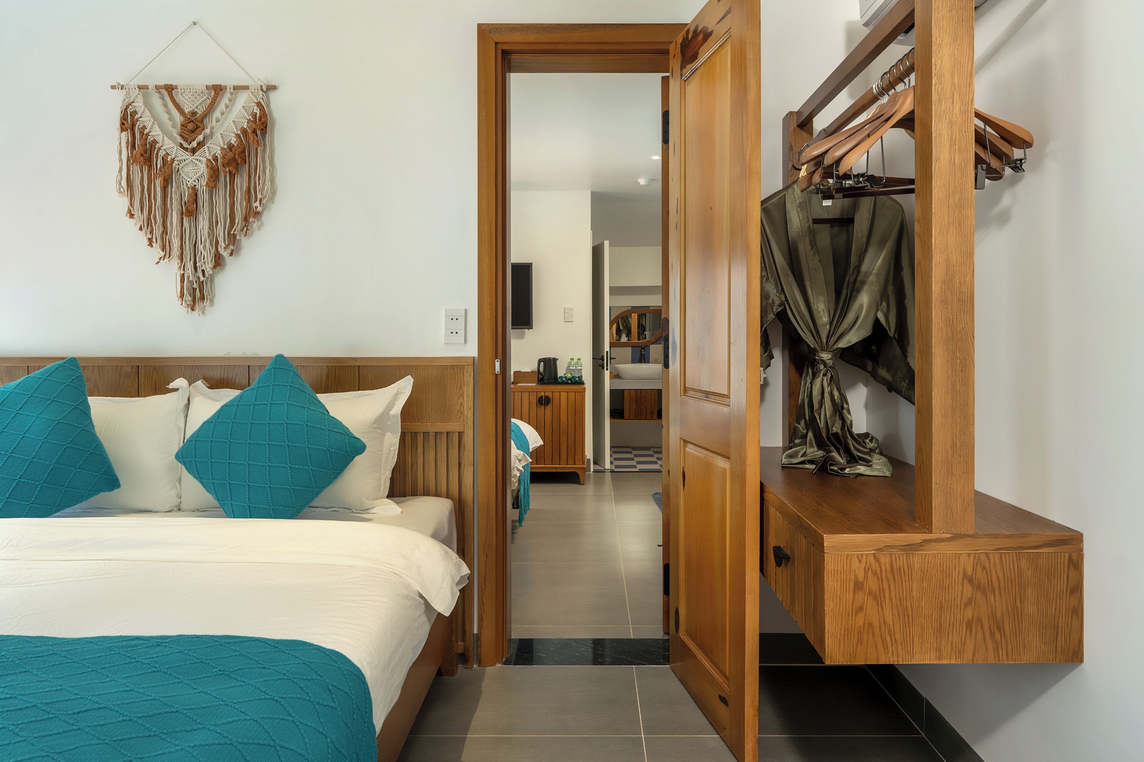 A hotel room with a bed, a wooden wardrobe, a hanging macrame wall decoration, and a view into the hallway.