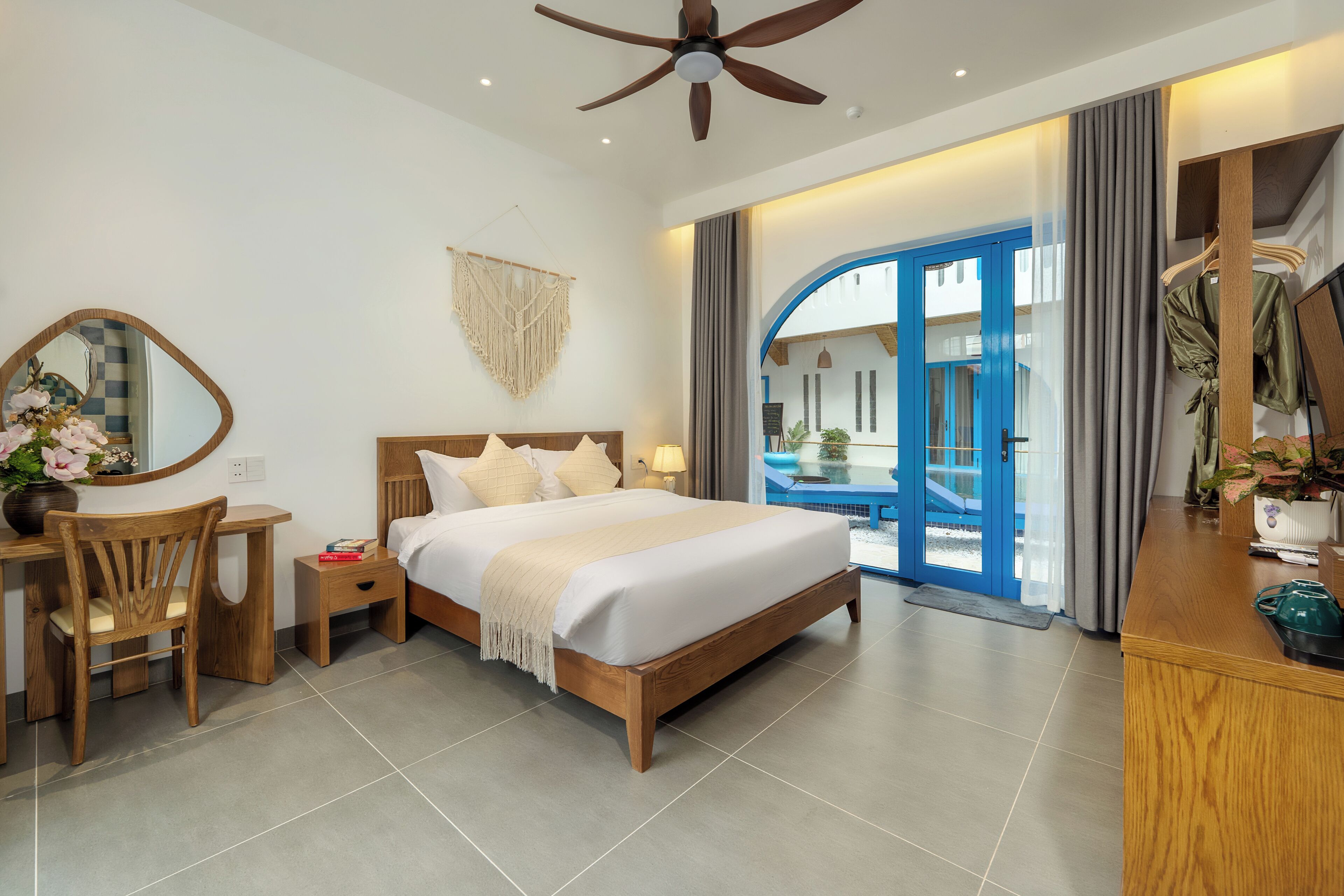 A modern hotel room with a wooden bed, a desk with a chair, a mirror, and a view of the outside through a glass door.
