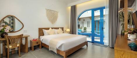 A modern hotel room with a wooden bed, a desk with a chair, a mirror, and a view of the outside through a glass door.
