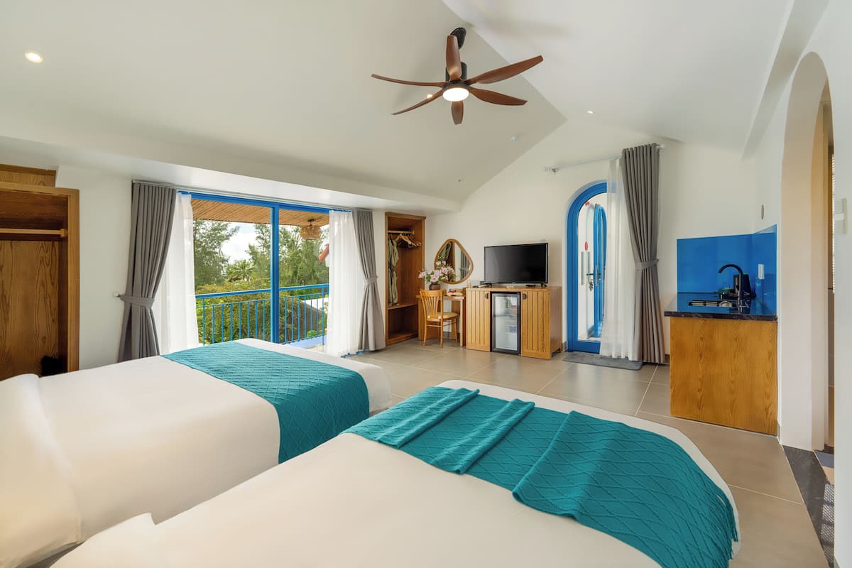 A hotel room with two beds, a ceiling fan, a TV, a small kitchenette, and a balcony with curtains.