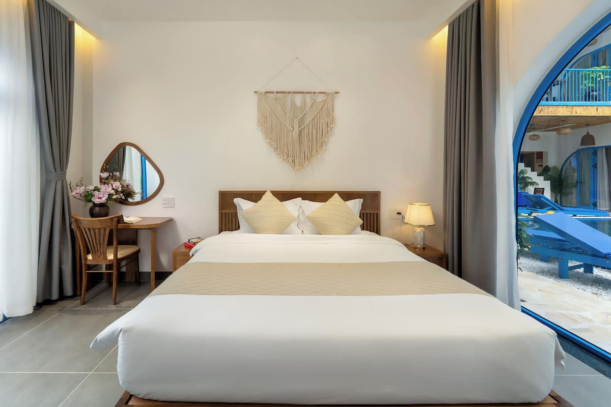 A hotel room with a large bed, a desk with a chair, a mirror, and a macrame wall hanging.