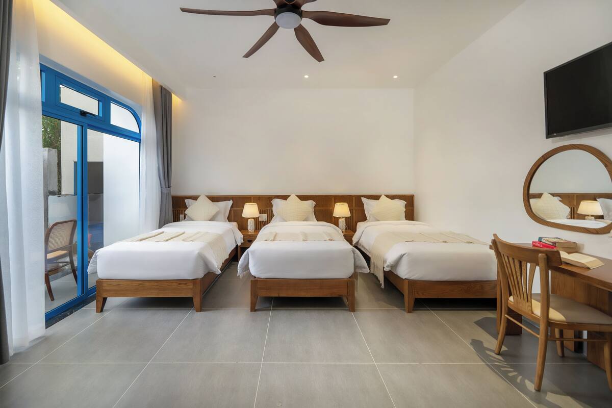 A hotel room with three beds, a ceiling fan, a TV, and a desk with a chair.