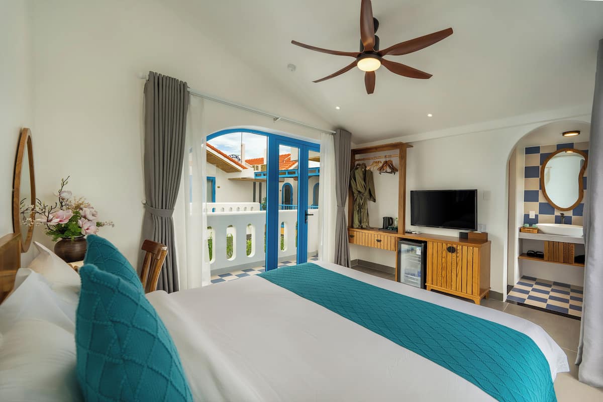 A hotel room with a bed, a desk with a TV, a bathroom with a mirror, and a balcony with a view of buildings.