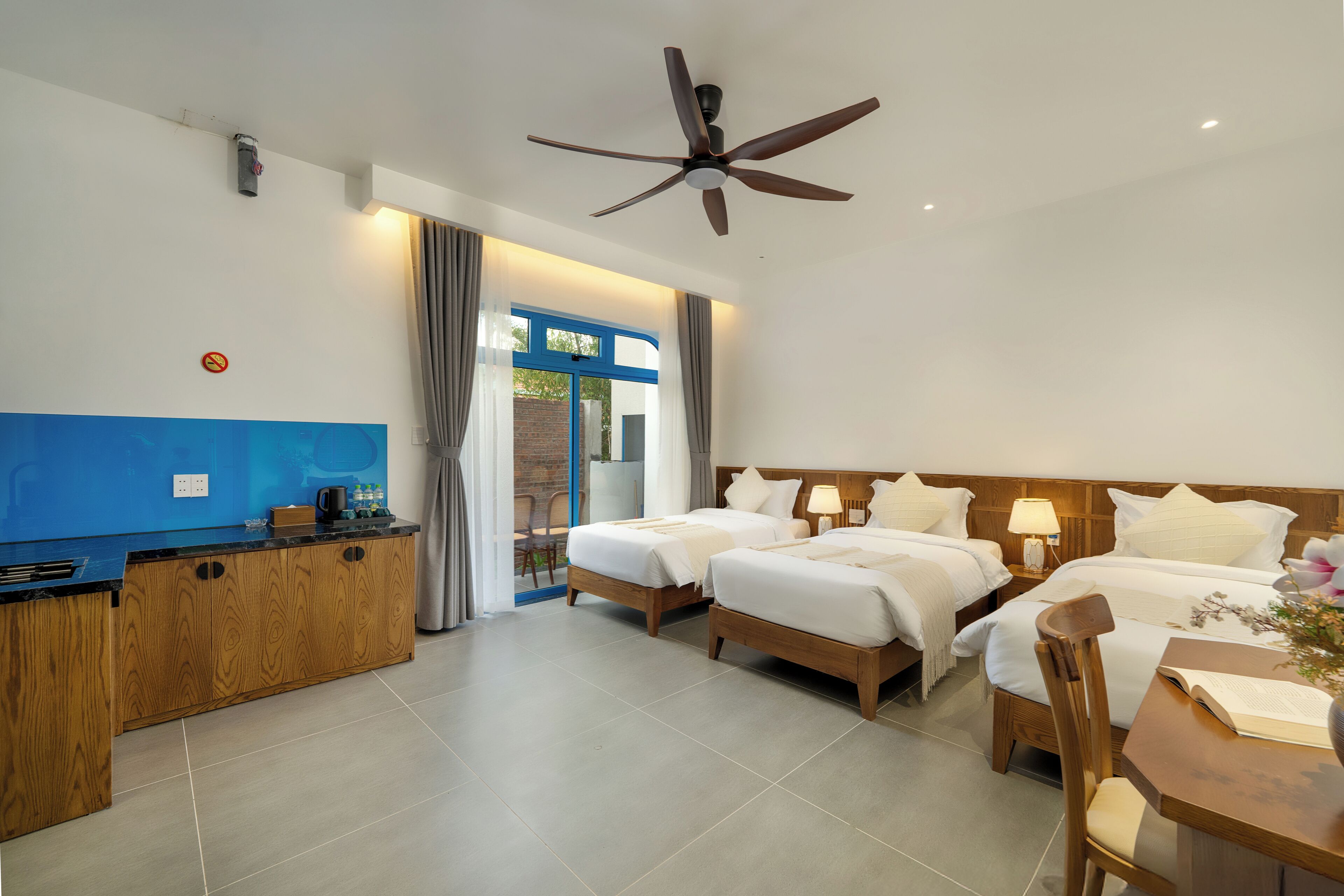 Quarto de hotel com duas camas, uma escrivaninha de madeira, um ventilador de teto e vista para o exterior.