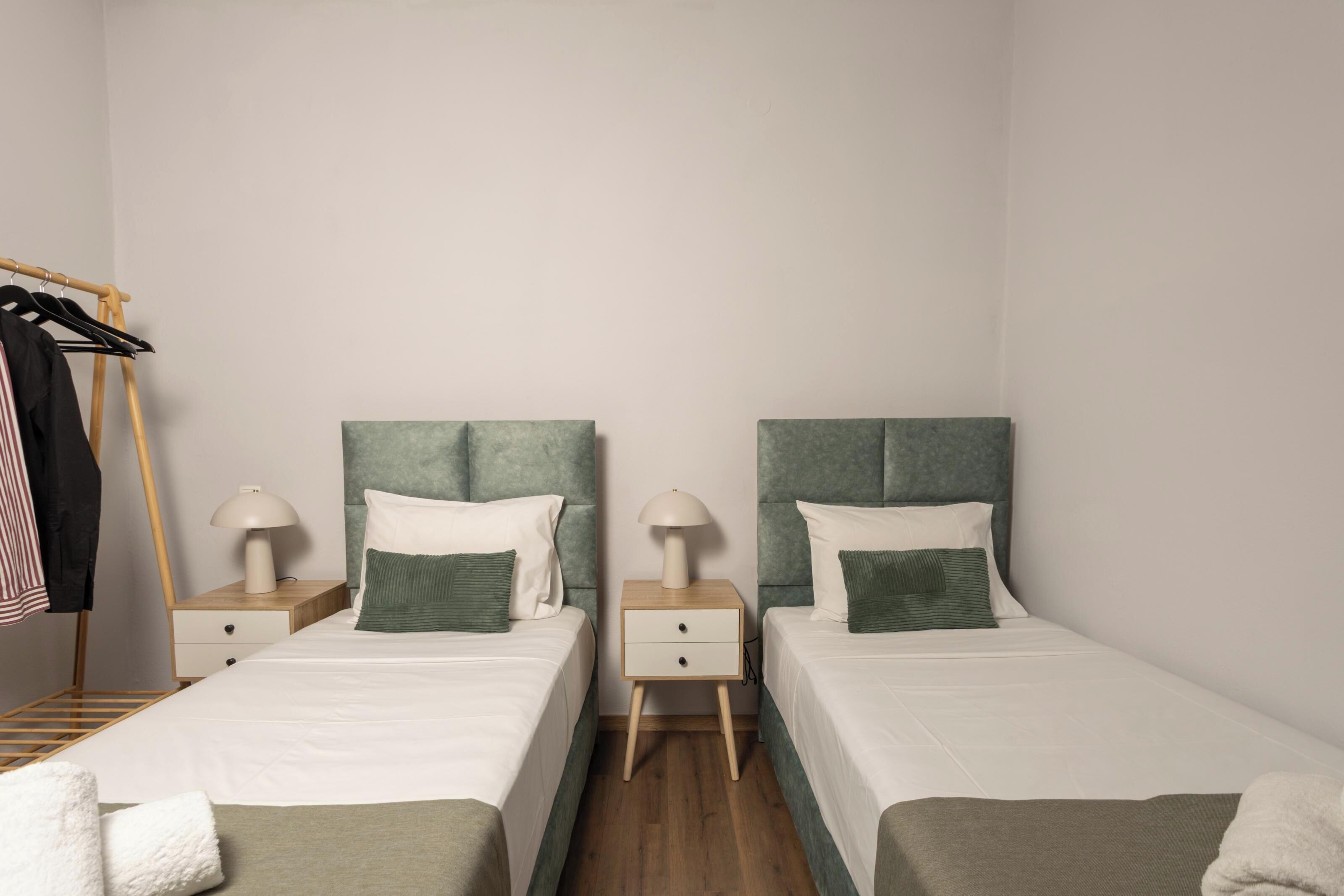 Habitación doble con dos camas, cada una con cabecero verde, ropa de cama blanca y una mesita de noche con lámpara. Se ve un armario de madera y un perchero.