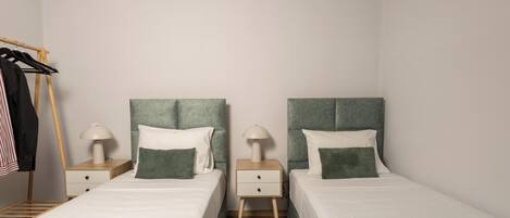 Habitación doble con dos camas, cada una con cabecero verde, ropa de cama blanca y una mesita de noche con lámpara. Se ve un armario de madera y un perchero.