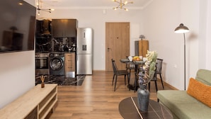 A modern kitchen with a refrigerator, oven, and washing machine. There is a dining area with a table and chairs, and a sofa with a lamp. - Zen City Apartment  (Chania)