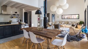 Une cuisine moderne avec une table en bois et des chaises blanches, un coin repas avec un canapé et une table basse, et une grande fenêtre qui laisse entrer la lumière naturelle.