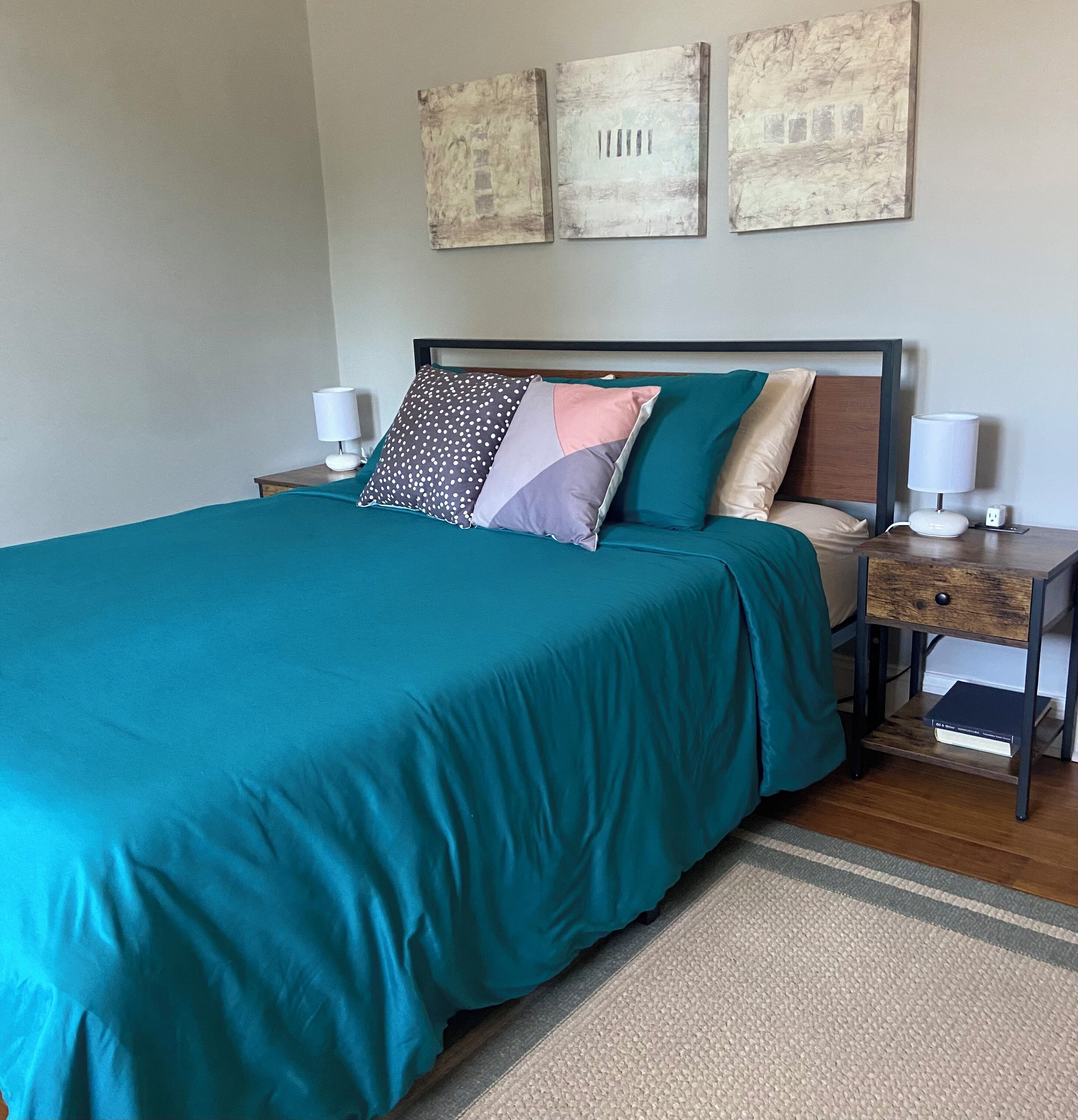 Una cama bien hecha con ropa de cama color turquesa, un cabecero de madera y dos lámparas de noche. Hay tres obras de arte colgadas en la pared sobre la cama.