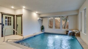 Une piscine intérieure aménagée comme un spa, avec une cascade intégrée, une porte et une fenêtre donnant sur la forêt.