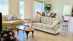 A living room with a beige sofa, a coffee table, and a dining area with a chair and a table. - Modern 4BR House Close to Beach & All Attractions (Palm Coast)