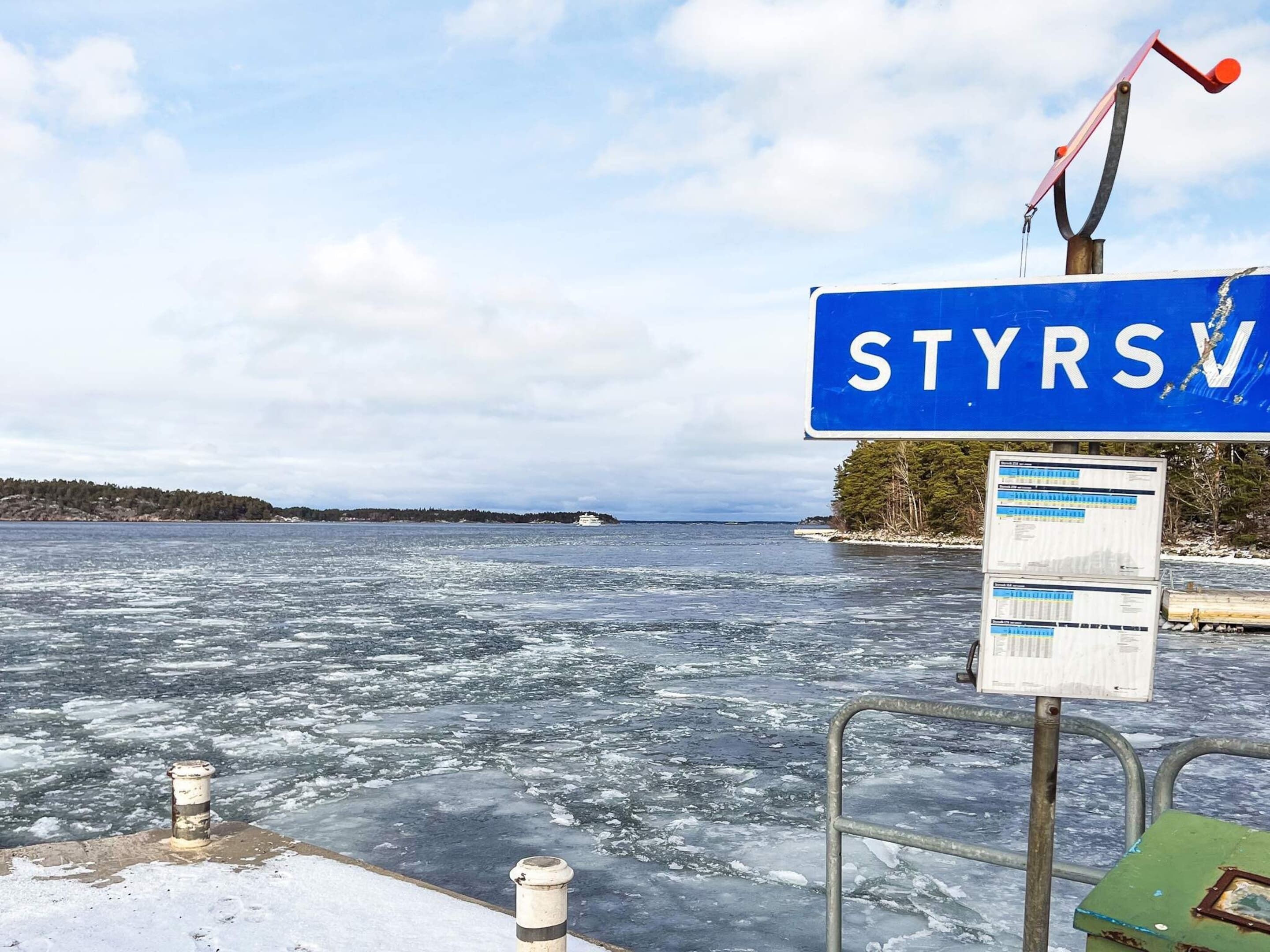 A sign with the text 'STYRSV' on a blue background, a metal railing, and a body of water with ice.