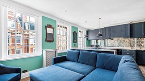 Un salon moderne avec un grand canapé bleu, un coin repas doté de meubles sombres et de grandes fenêtres offrant une vue sur la ville.