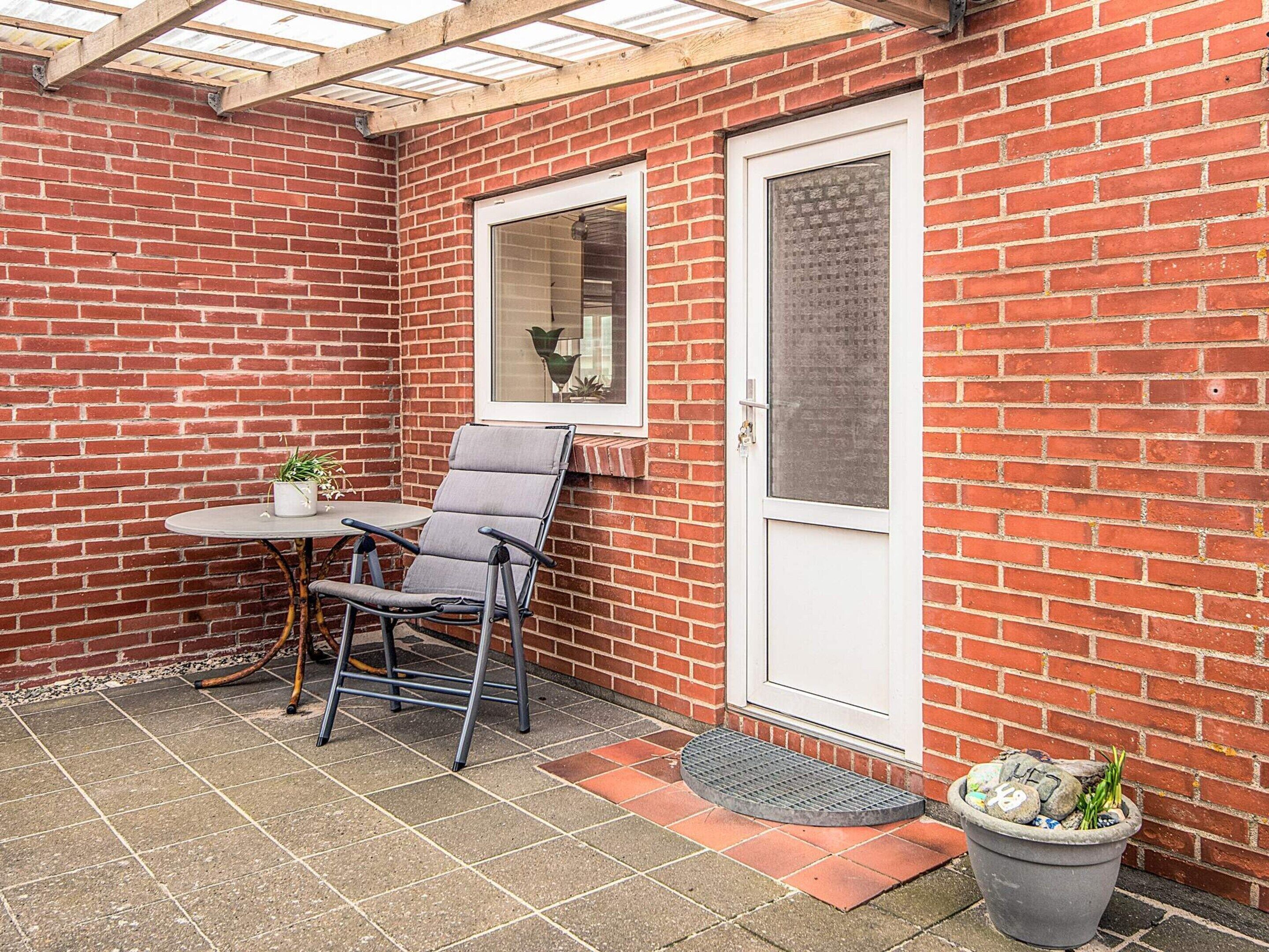 A small patio with a brick wall, a white door, a window, a table with a chair, and a potted plant.