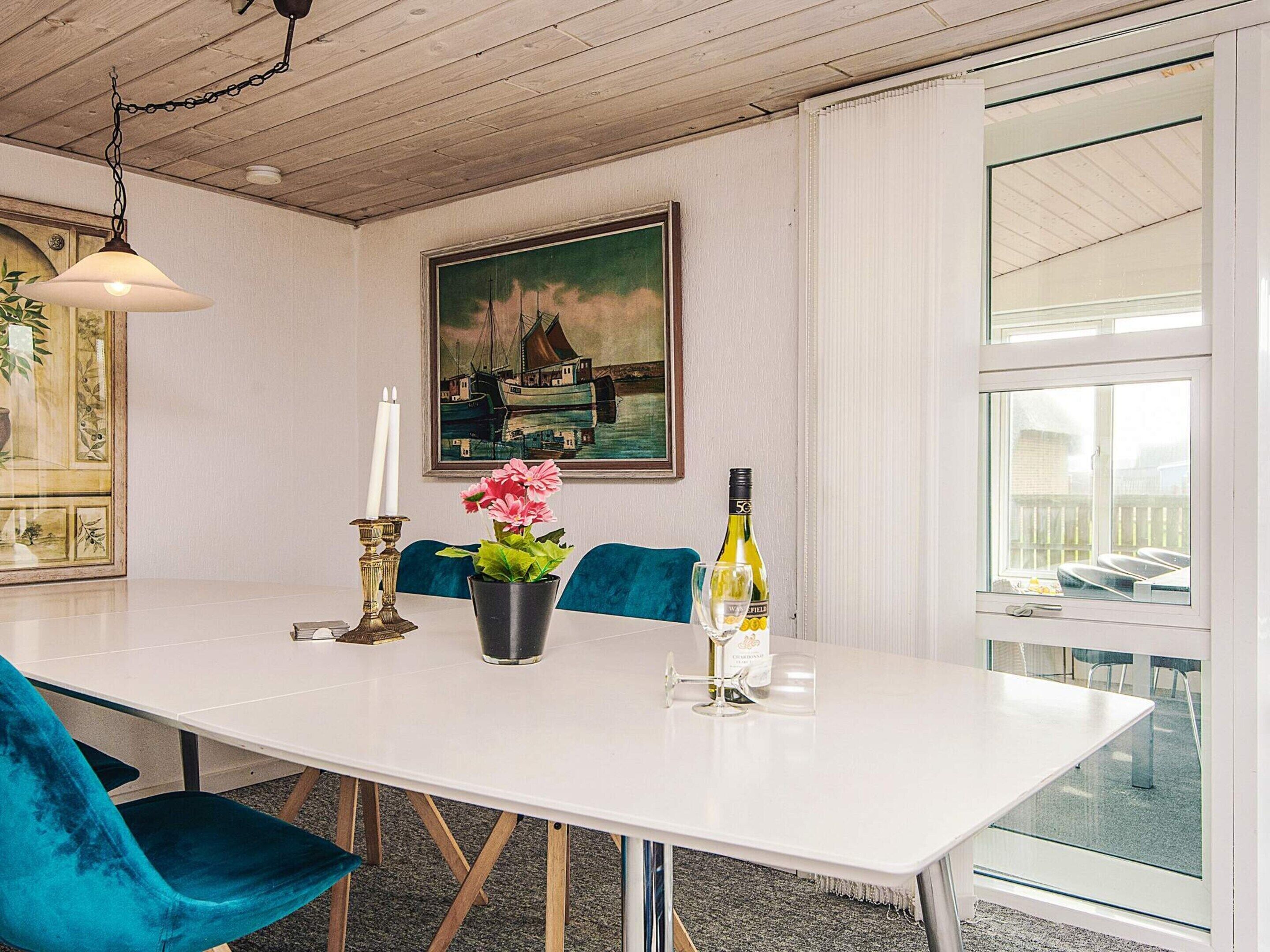 A dining area with a white table, blue chairs, a painting of a harbor scene, a candle, a flower vase, and a wine bottle.