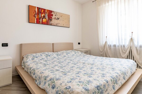 Une chambre à coucher comprenant un lit, une table de chevet, une commode, une œuvre d’art accrochée au mur et une fenêtre ornée de rideaux légers.