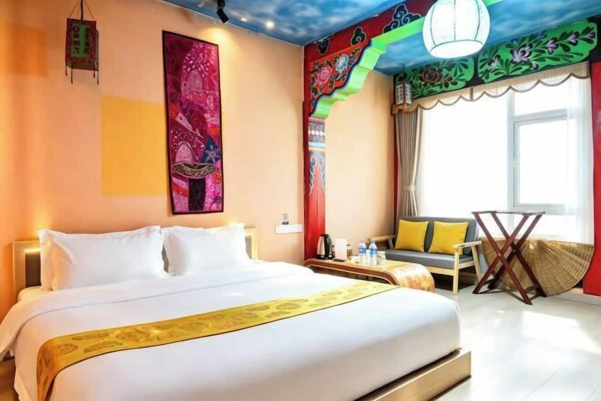 Ein Hotelzimmer mit einem großen Bett, einer Sitzecke mit Stuhl und Tisch, einem großen Fenster mit Vorhängen sowie einer farbenfrohen Wanddekoration.