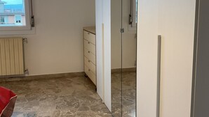 A room with a tiled floor, a window with blinds, a radiator, a wardrobe, and a mirror.