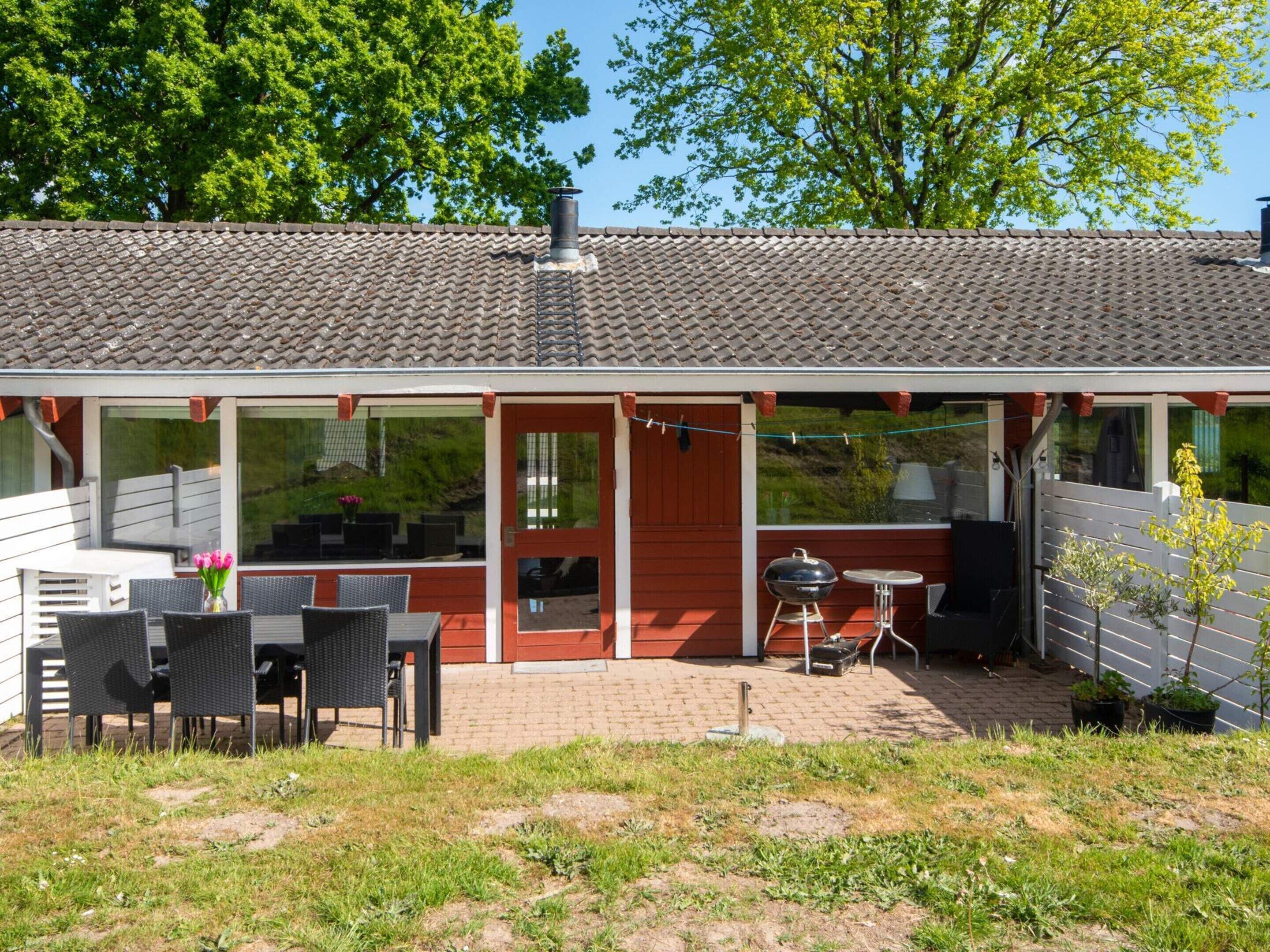 A small, single-story building with a tiled roof, a red door, and a patio area with outdoor furniture.