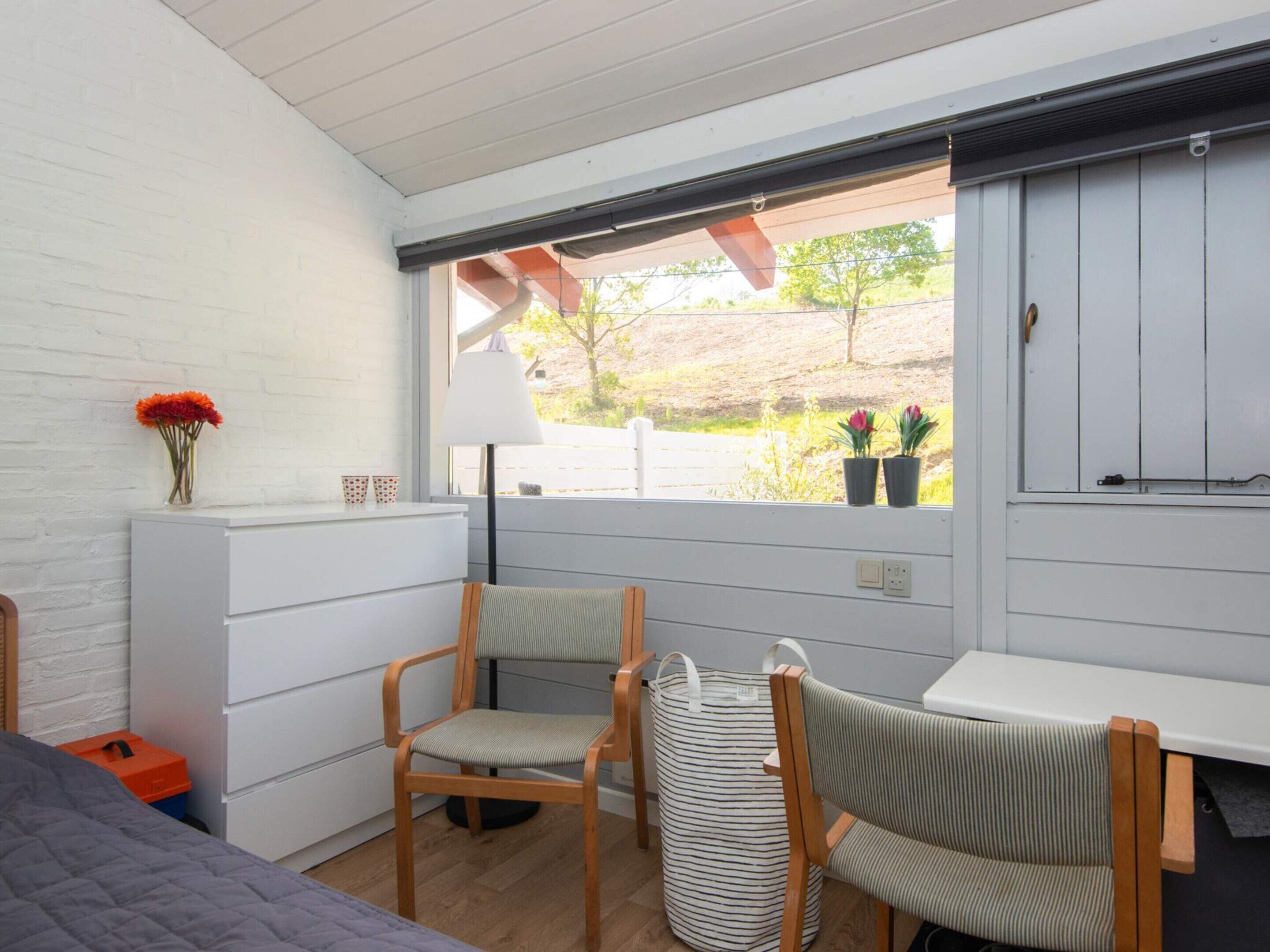 A room with a bed, a chair, a table, a window with a view of greenery, and a vase with flowers.