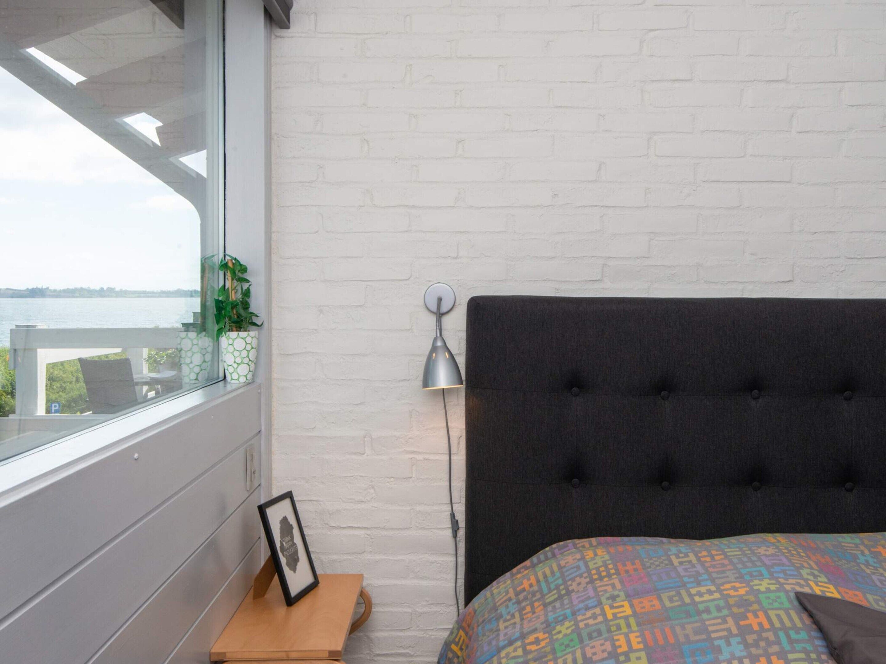 Un dormitorio con pared de ladrillo blanco, un ventanal con vista a una masa de agua, una cama con colcha estampada, una mesita de noche con lámpara y un cuadro enmarcado.