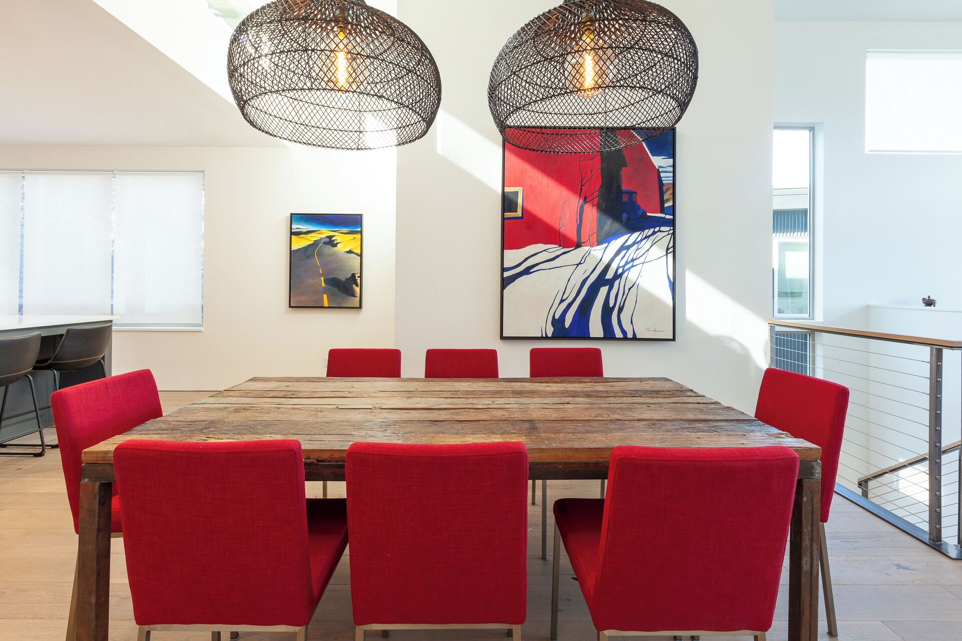 A modern dining area with a wooden table and red upholstered chairs, two hanging pendant lights, and two large abstract paintings on the walls.