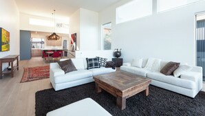 A spacious living room with a white sectional sofa, a wooden coffee table, and a black rug. There is a kitchen in the background with red chairs and a hanging light fixture. - NEW Construction, Magnificent NEW Townhome in dt Hood River w/Rivr views, garage (Hood River)