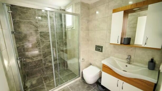 A modern bathroom with a glass-enclosed shower, a white toilet, and a vanity with a mirror and sink. - Aydınsuit (Izmir)