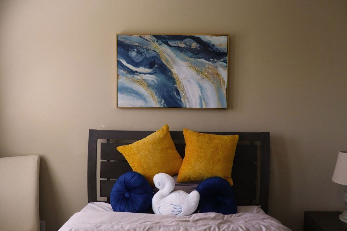 Een bed met een witte dekbedovertrek, twee gele kussens en een blauw kussen. Daarboven hangt een schilderij van abstracte vormen in blauw en wit.