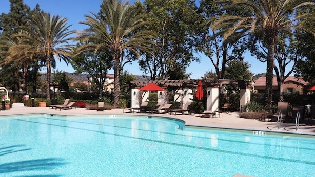 A clear swimming pool with lounge chairs and umbrellas, surrounded by palm trees and a building with a red roof. - Pretty Relaxed Me: Pool, Spa, Clubhouse (Brea)