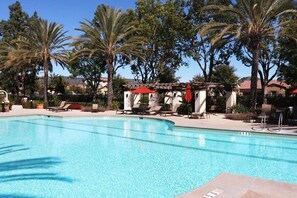 A clear swimming pool with lounge chairs and umbrellas, surrounded by palm trees and a building with a red roof.