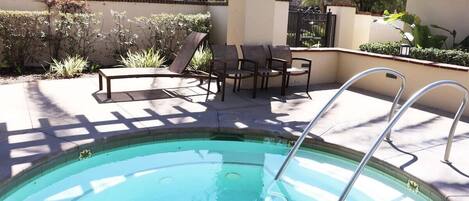 A swimming pool with a sun lounger and chairs, surrounded by a tiled deck and a low wall with plants.